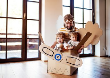 Ein Erzieher sitzt mit zwei Kindern in einem selbstgebauten Flugzeug aus Pappe
