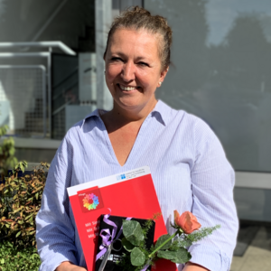 Zerina Music-Neuhaus, Absolventin der Weiterbildung Praxisanleitung, steht lächelnd vor einem Gebäude. Sie hat eine rote Rose und eine rote Mappe in der Hand.